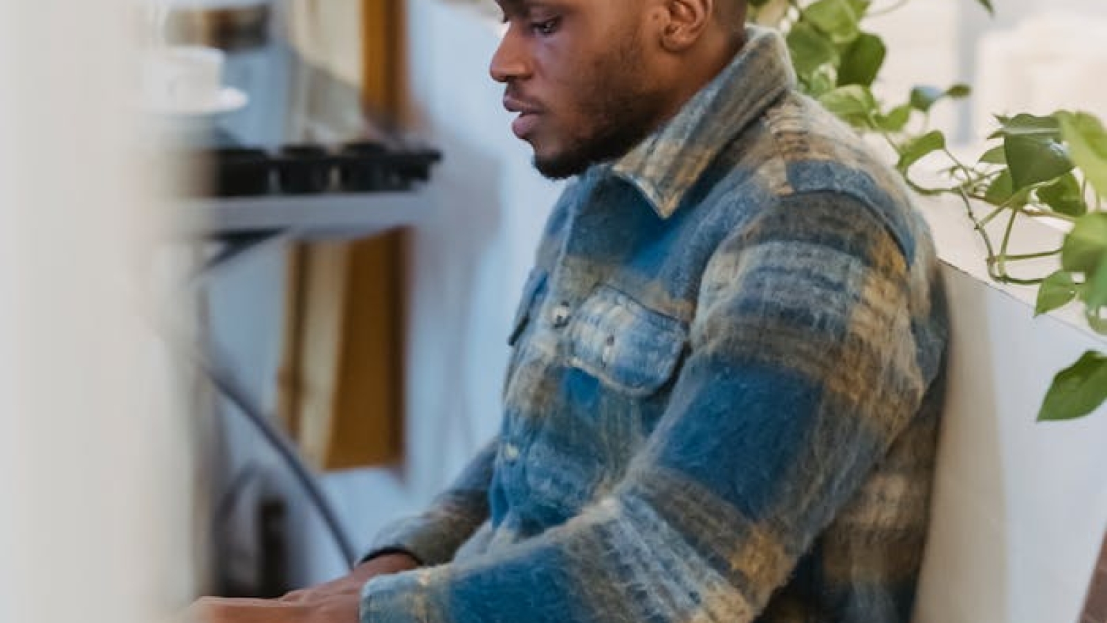 Side view of focused bearded African American guy in checkered shirt sitting near white partition decorated with green plant and working remotely on portable computer