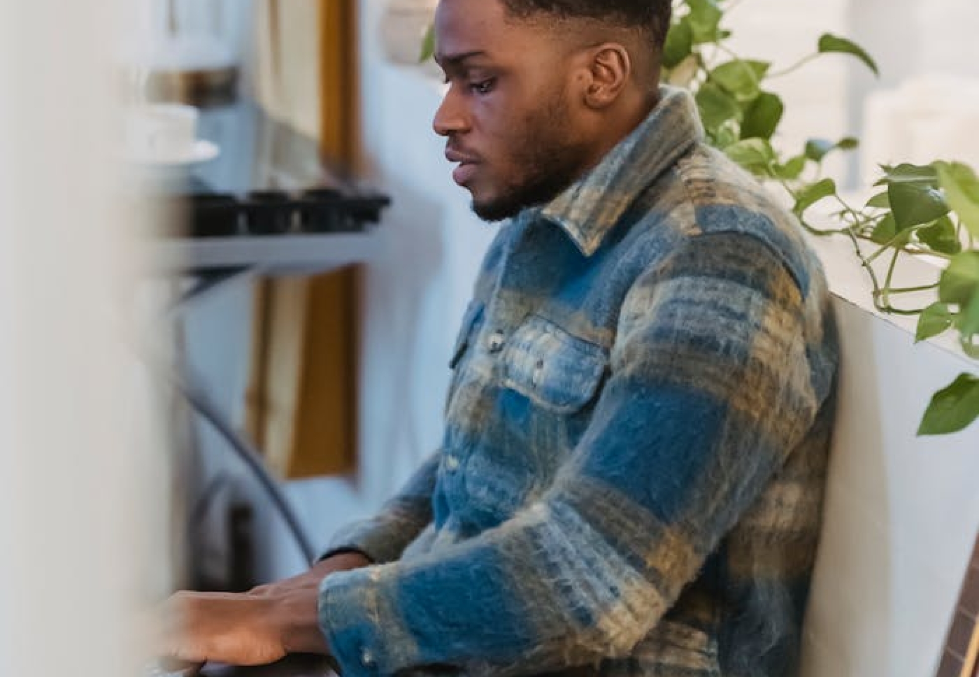 Side view of focused bearded African American guy in checkered shirt sitting near white partition decorated with green plant and working remotely on portable computer