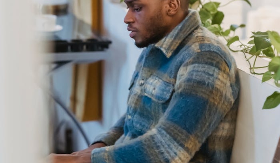 Side view of focused bearded African American guy in checkered shirt sitting near white partition decorated with green plant and working remotely on portable computer