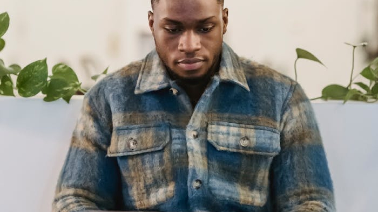Concentrated young African American male freelancer in checkered shirt typing on laptop keyboard while working remotely in cafe