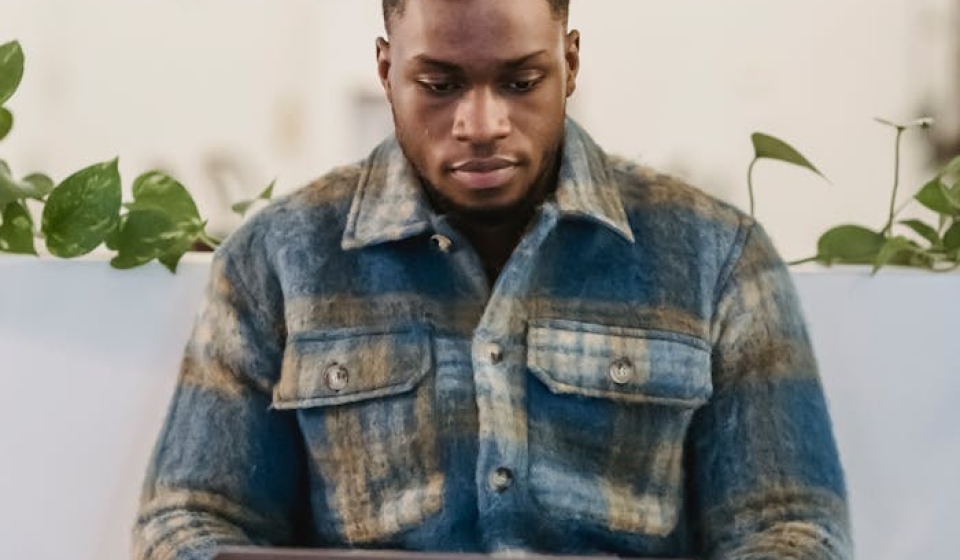 Concentrated young African American male freelancer in checkered shirt typing on laptop keyboard while working remotely in cafe